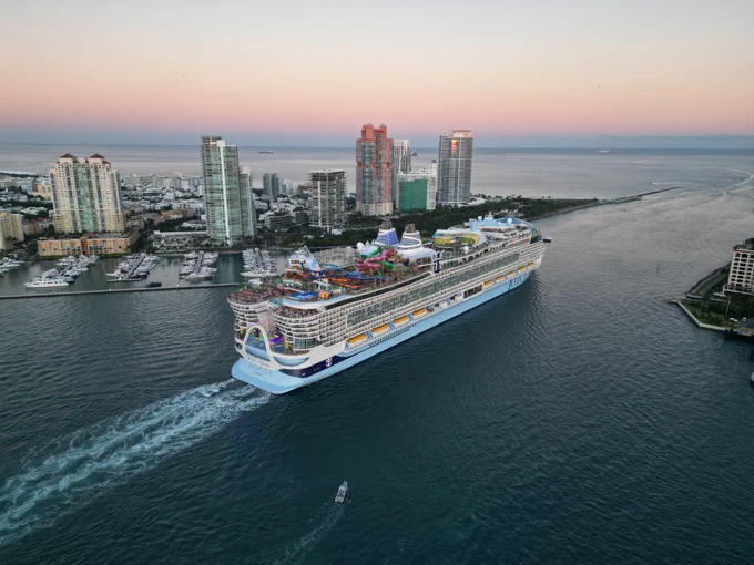 Aerial view of the Icon of the Seas cruise ship sailing on the ocean with the Miami skyline in the background at sunset. The ship has water slides and many decks. A small boat follows behind the cruise ship.