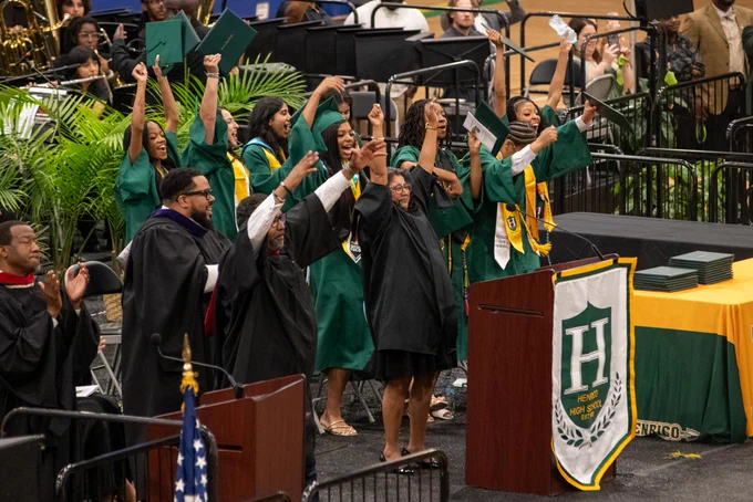 A group of people in green and yellow graduation gowns and caps celebrating at a graduation ceremony, with a stage and backdrop visible.