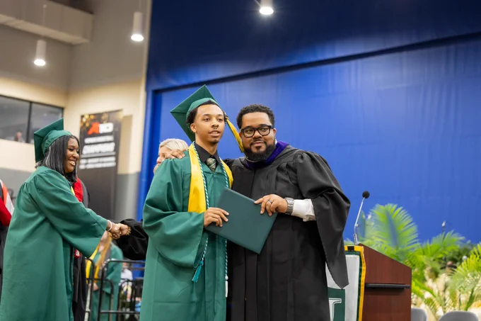 Two graduates in green and black gowns standing together, smiling and holding diplomas.
