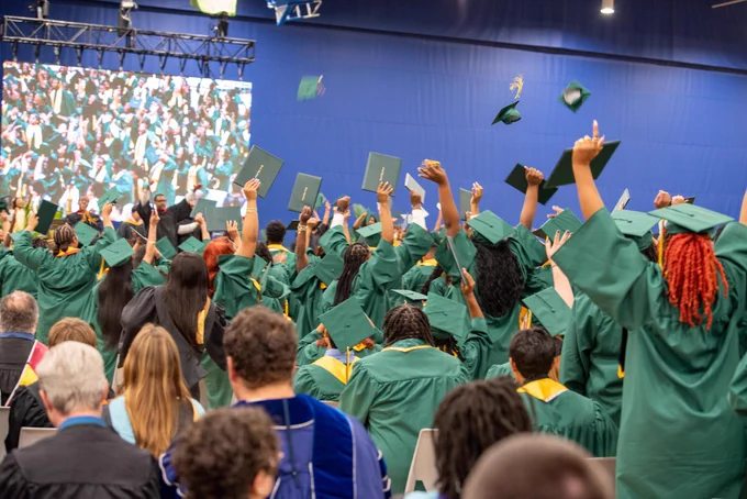 Graduation ceremony with a crowd of students in green gowns and mortarboards throwing their hats into the air.