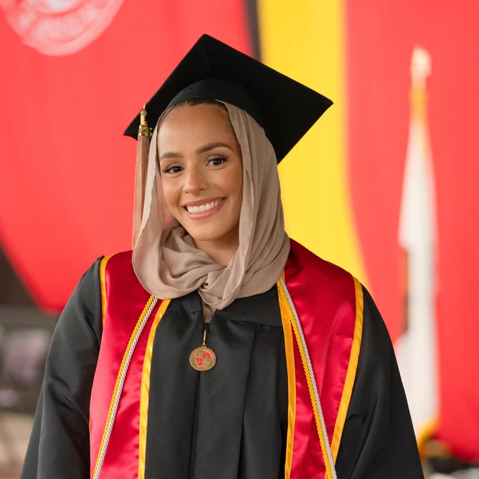 A smiling graduate wearing a hijab and graduation cap poses confidently in a black gown with red and gold Stanislaus State sash. Celebratory atmosphere.