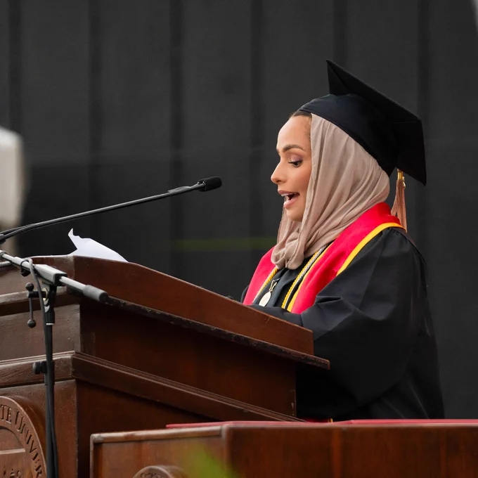 A woman in a graduation gown delivers a speech at a graduation ceremony, standing confidently at a podium.