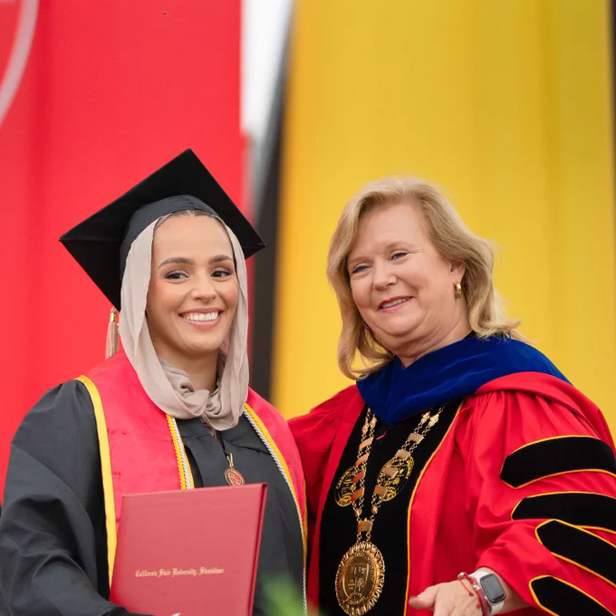 Graduate in cap and gown smiles holding diploma, standing beside a smiling faculty member in academic regalia. Bright banners in background. Graduation ceremony.