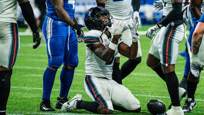 A football player in a white uniform kneels on the field with hands raised, celebrating. Several players in blue and white uniforms surround him on the grass.