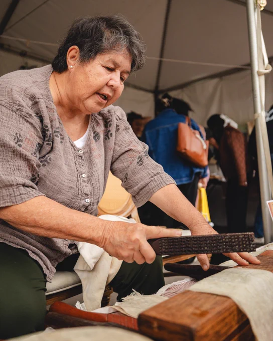 A person seated under a tent is demonstrating traditional weaving using wooden tools on a wooden frame. The setting appears to be a cultural or craft event.