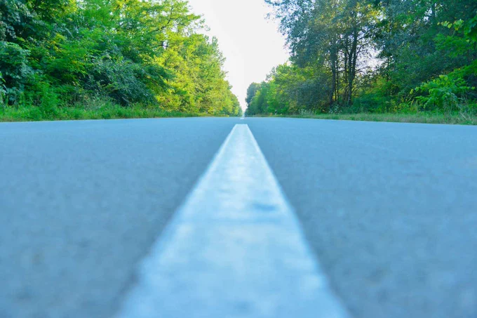 A straight road with a single white center line stretches into the distance, surrounded by lush green trees on both sides under a clear blue sky.