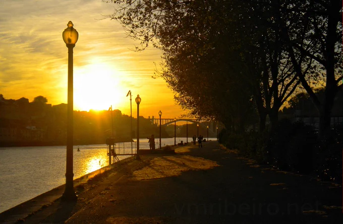 Passeio ribeirinho ao pôr do sol, com árvores a projetar sombras, pessoas a caminhar, candeeiros acesos e a ponte da Arrábida visível ao fundo sobre o rio Douro.