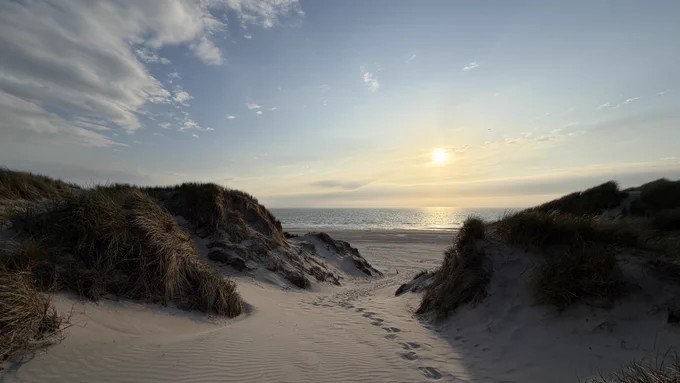 Blick über Dünen in Richtung Strand, Meer und Horizont mit einem mittigen Abgang zum Strand. Die Sonne geht bald unter.
