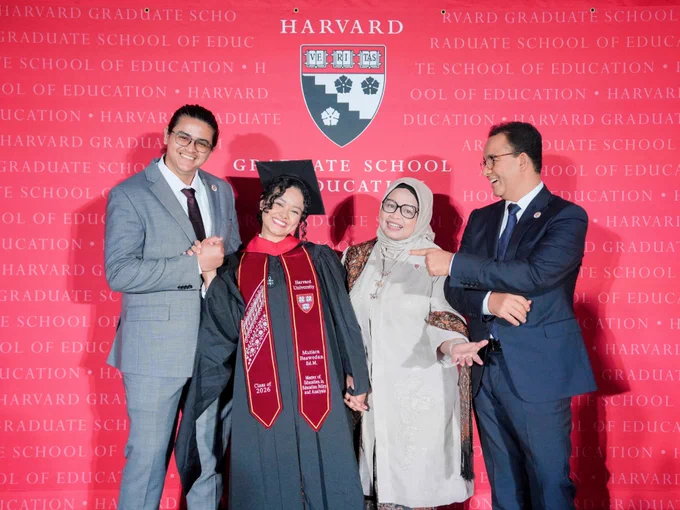 Mutiara Baswedan tersenyum mengenakan toga hitam, mortarboard, dan selempang merah Harvard dalam acara wisuda Harvard Graduate School of Education. Tia berdiri di antara Ali Alhuraiby (suaminya), Fery Farhati, dan Anies Baswedan, yang tampak tersenyum bangga sambil menunjuk ke arah Tia. Mereka berpose di depan latar merah bertuliskan “Harvard Graduate School of Education” dengan lambang Harvard di bagian tengah atas.