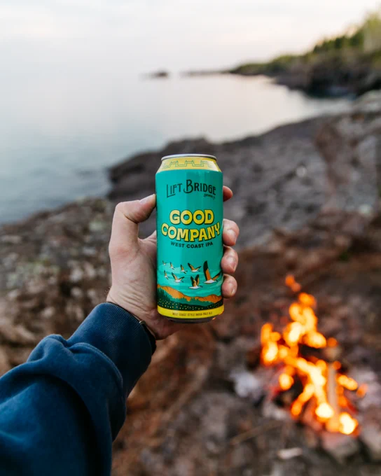 A vibrant can of West Coast IPA held in focus by a campfire, with a serene lakeside backdrop. The image conveys a relaxed outdoor vibe.