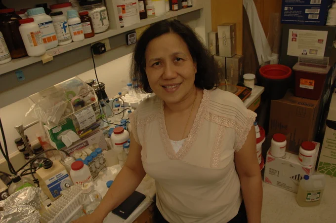 Nimman Satumtira wearing a tan blouse. She is standing in front of a lab table and cabinet covered in different bottles, containers, and scientific equipment.