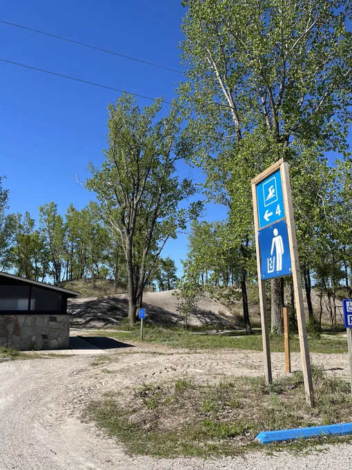 A blue sign pointing to the beach and a garbage station with trees, sand dunes, and a comfort station. 