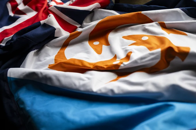 Blue flag with Union Jack and a crest of three orange dolphins in a circle on a white background.