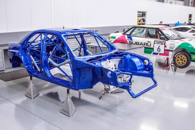 Subaru Impreza WRC bodyshell during restoration at Prodrive Legends workshop in Banbury, showing roll cage structure before rebuild to 2004 Rally Japan spec. Behind it in the next bay is Ari Vatnen’s 1992 Subaru Legacy