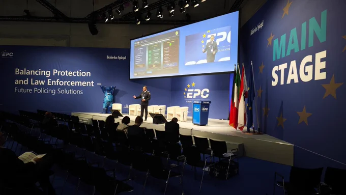 Escenario principal con presentador frente a gran pantalla; conferencia sobre equilibrar protección y aplicación de la ley.