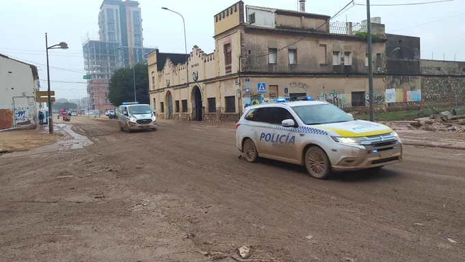 Coches de policía con luces azules circulan por una calle embarrada frente a un edificio antiguo con grafitis.