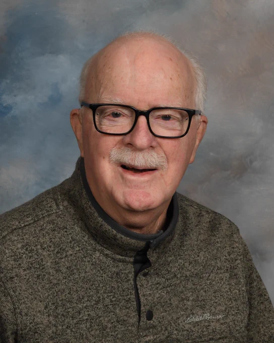 An elderly man with glasses, light skin, and a white mustache smiles softly. He wears a gray sweater against a cloudy, neutral-toned background.