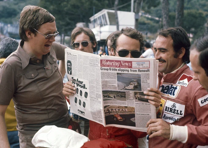 Niki Lauda, wearing aviator sunglasses in the centre of frame, holds up a copy of the Motoring News  as he reads it. On the right of frame, Clay Regazzoni smiles as he too looks at the pages; Laughing on the left of frame, Alan Henry stands in a brown polo shirt and sunglasses.