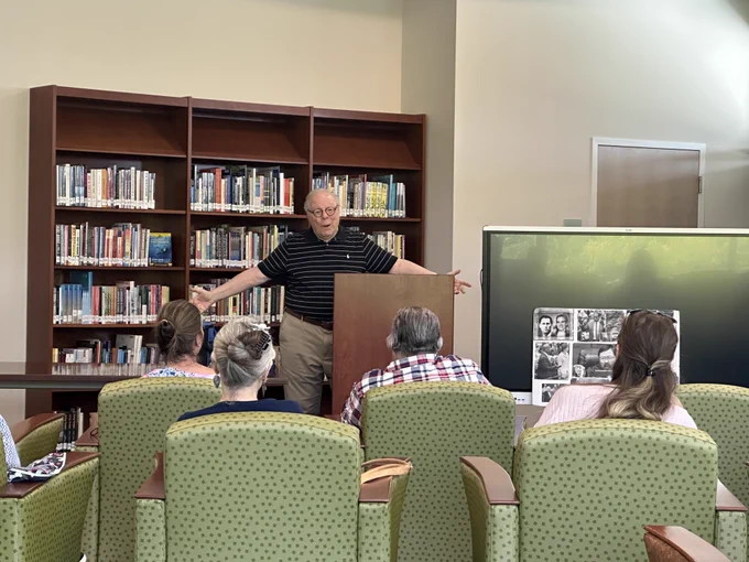 A man stands behind a wooden podium speaking to a small audience seated in green upholstered chairs in a library setting, with bookshelves filled with books behind him and a display board featuring black-and-white photographs to his right.
