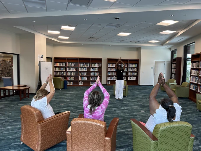 A group of four people practicing seated yoga with arms raised in a library setting surrounded by bookshelves and comfortable chairs.