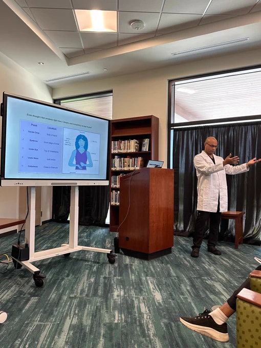 A man in a white lab coat giving a presentation in a room with a large screen displaying a slide about health topics, with an audience seated nearby.