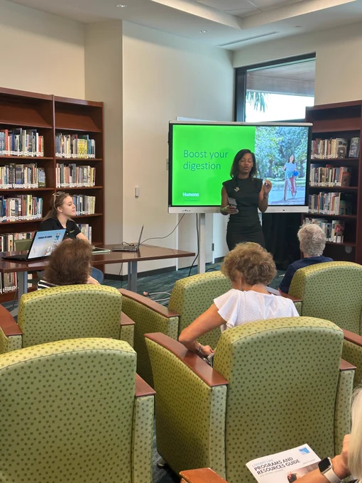 A presenter stands near a screen displaying 'Boost your digestion' while an audience seated in green chairs listens attentively in a library setting.