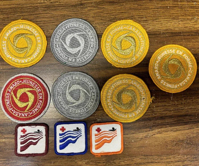 Nine embroidered fitness award patches are arranged on a scratched wooden tabletop. Six larger circular patches across the top and middle rows have yellow, silver, red, and gold designs with the Canada Fitness Award text in English and French around the edge. Three smaller square swimming patches sit along the bottom, bordered in maroon, blue, and orange with wavy lines and a red maple leaf.
