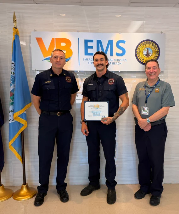 Three people in uniform stand in front of a sign reading "VB EMS" and a city seal. The person in the middle holds a certificate. A flag is visible to the left.