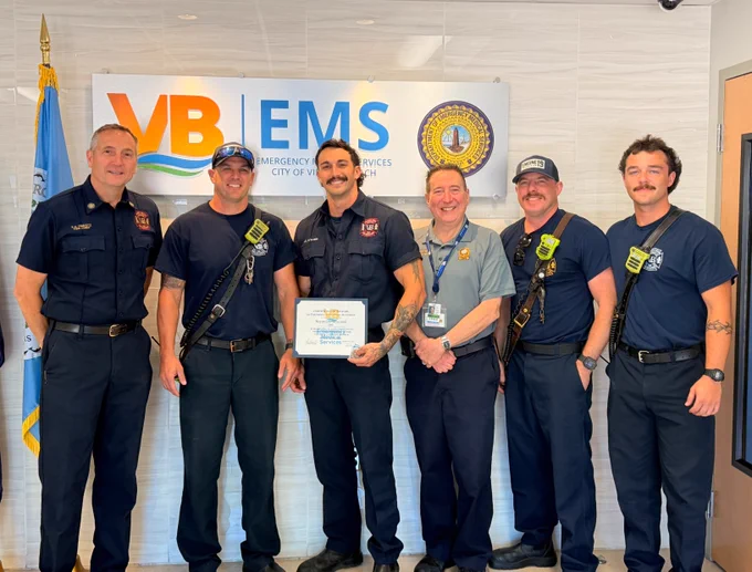 A group of six people in uniform stand together smiling, with one holding a certificate. They are in front of a sign that reads "VB EMS Emergency Medical Services City of VB." An official emblem is displayed on the wall.