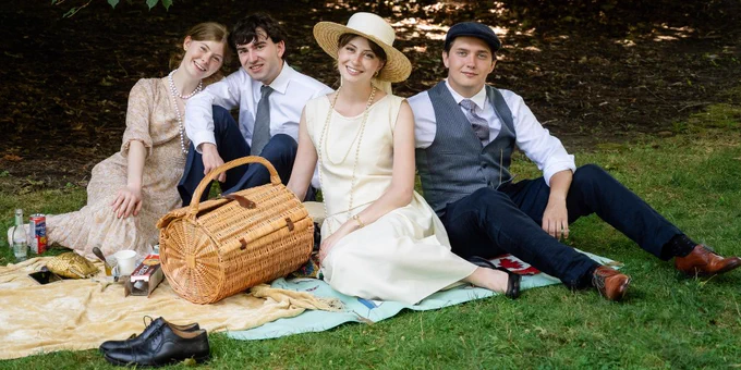 Four friends dressed in vintage attire enjoying a picnic on a blanket under the shade of trees in a grassy park.