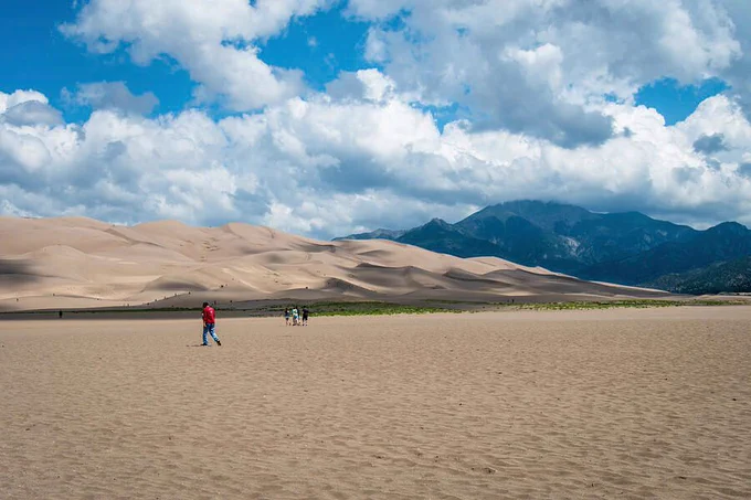 The Great Sand Dunes National Park in Colorado has the tallest dunes in North America, with the legendary Star Dune towering at 755 feet. On this day, beautiful white puffy clouds float over the landscape of dunes and mountains.  Photography by Debra Martz