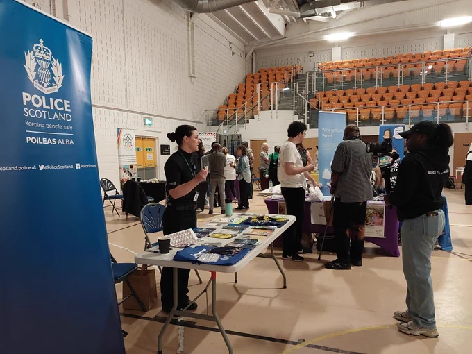 Police Scotland officer at a community event, with stalls and attendees in the background.