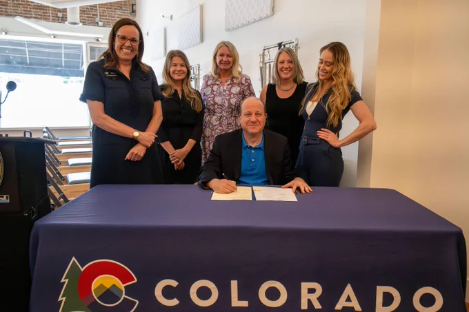 Group photo of Governor Jared Polis signing a bill at a table covered with a Colorado logo, surrounded by five standing women indoors.
