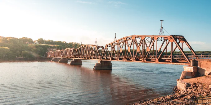 Wide view of a rust-colored railroad truss bridge spanning a river in warm sunlight, with stone bridge piers, wooded bluffs, rippling water, and a rocky shoreline in the foreground.