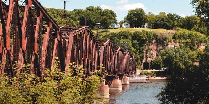 Side view of a long rust-colored steel railroad truss bridge crossing a river, with wooded bluffs, exposed rock faces, and houses on the hilltop in the background.