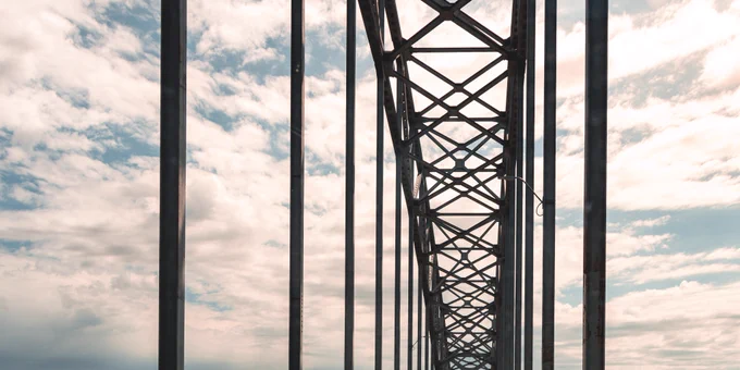 Looking upward through the vertical steel members and overhead cross-bracing of the Julien Dubuque Bridge, with a bright partly cloudy sky visible behind the dark structural frame.