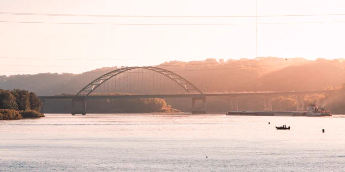 Warm, hazy view of an arched highway bridge crossing a wide river at sunset, with wooded bluffs in the background, a barge and tugboat on the water, and a small fishing boat in the foreground.