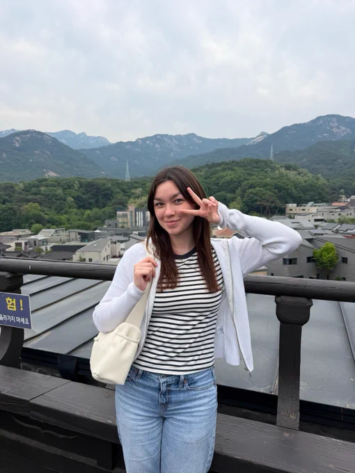 A person stands on a rooftop with a scenic view of mountains and greenery in the background. They are wearing a white cardigan, striped shirt, and jeans, holding a white bag. The person is making a peace sign with one hand. It's a cloudy day.