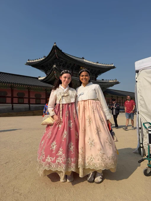 Two people wearing traditional Korean hanboks stand smiling in front of a historic building at Gyeongbokgung Palace. The hanboks are decorated with intricate embroidery. Other visitors are seen in the background under a clear sky.