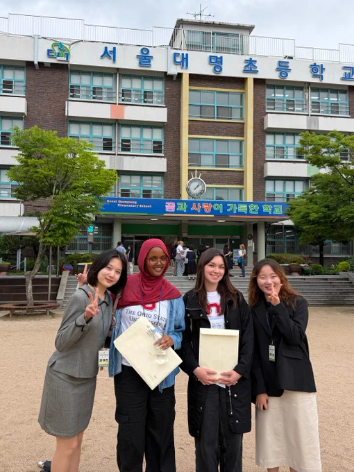 A group of four individuals stands in front of a building with a sign that reads "서울대명초등학교." Two people in the center hold certificates. The building features a clock above the entrance and Korean text in blue and green. The individuals appear to be smiling and gesturing towards the camera.