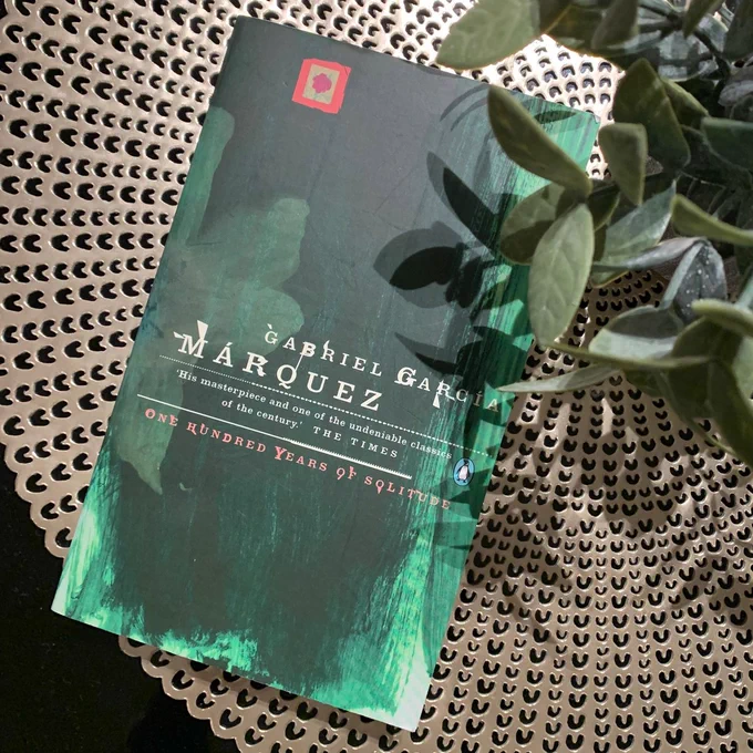A hardcover book titled 'One Hundred Years of Solitude' by Gabriel García Márquez, lying on a tabletop, partially covered by green plant leaves.