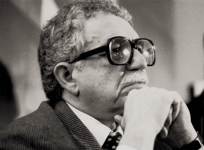 Black and white photo of Gabriel García Márquez in thoughtful pose, resting chin on hand, wearing glasses and a suit.