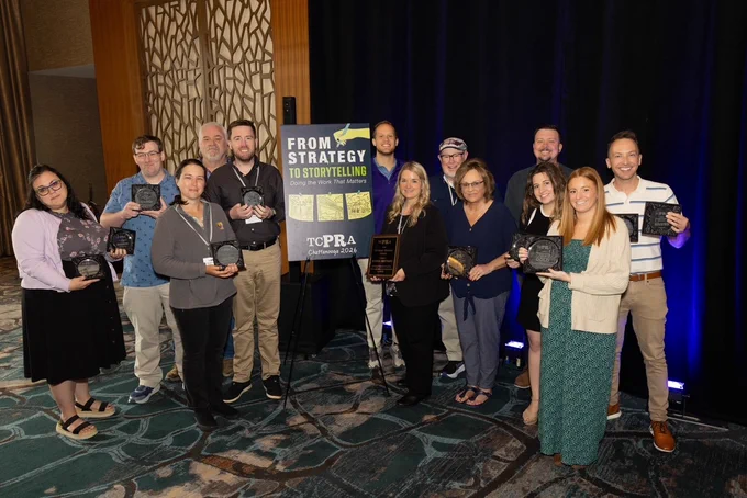A group of thirteen people stands in a room, holding awards and smiling. They are positioned around a sign that reads "From Strategy to Storytelling, TCPRA Chattanooga 2026." The setting appears to be an award ceremony with a patterned carpet and a decorative backdrop.