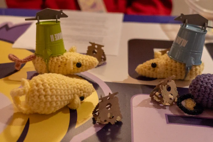 Three crochet Ratales on a table together, two with buckets on their head, surrounded by miniature cardboard cheese pieces