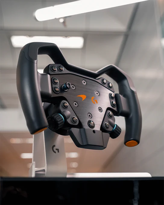 Volante de RS Formula Wheel McLaren Racing Edition, mayormente negro con partes naranjas. Está ubicado en una habitación moderna y bien iluminada.