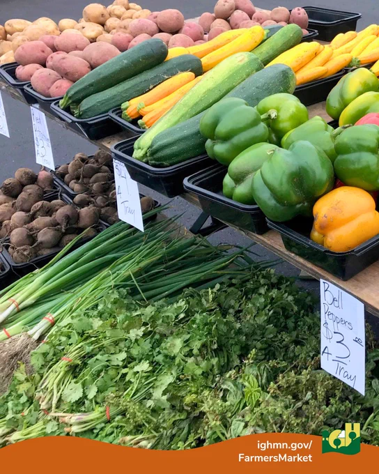 Fresh green and yellow zucchinis, bell peppers, potatoes, and leafy herbs displayed on trays at a farmers market stand.
