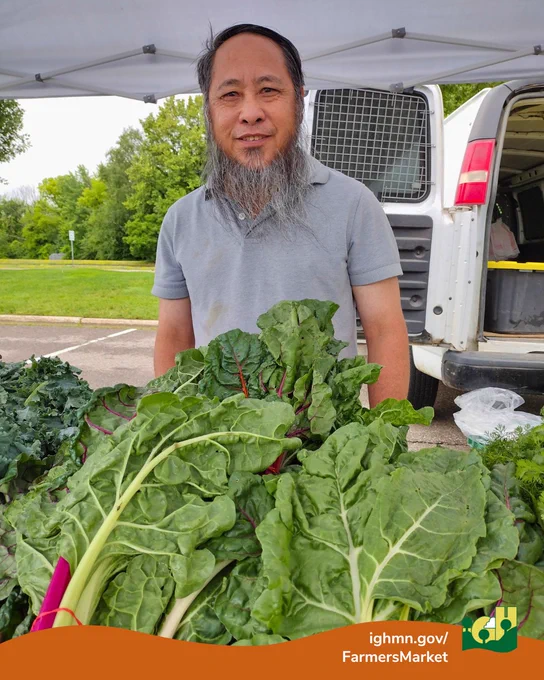 Vendor standing behind a table full of fresh leafy greens at an outdoor farmers market with a white van in the background.