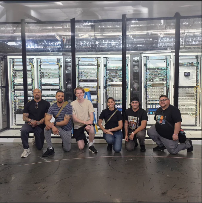 A group of six people posing in front of a large rack of Cisco networking equipment.