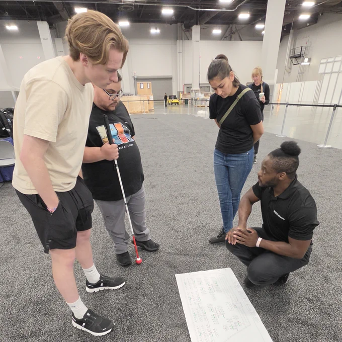 Four people in an event hall reviewing a large technical diagram on the floor, with one individual using a white cane.