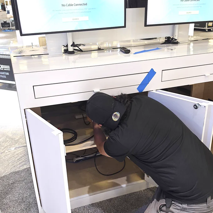 A person kneeling and organizing cables inside the cabinet of a white display desk with two monitors.